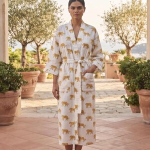 Könnte beinhalten: Ein wadenlanger Bademantel mit langen Ärmeln in Off-White, der mit einem sich wiederholenden Muster aus gelben und braunen Leoparden bedruckt ist. Der Mantel verfügt über einen passenden Bindegürtel, einen Schalkragen und zwei aufgesetzte Taschen.
