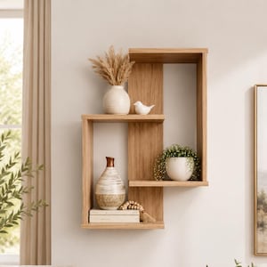 May include: A light oak wood wall shelf with an asymmetrical geometric design featuring multiple open compartments. The shelf holds a white textured vase with dried pampas grass, a small white bird figurine, a ceramic vase, and a potted plant.