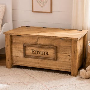 May include: A rectangular wooden toy chest with a natural light brown finish and visible wood grain. The front panel features a centred rectangular plaque engraved with the name Emma in a dark serif font. The chest has a flat hinged lid.