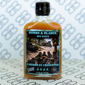 May include: A glass bottle of Bombs & Blades Caribbean Crosswind hot sauce with a black cap. The label features a military-themed graphic, the text 'BOMBS & BLADES HOT SAUCE', 'CARIBBEAN CROSSWIND', and '6.8 FL OZ (201ML)' with four flame icons.