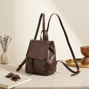 Può includere: Uno zaino in pelle marrone scuro con patta ripiegabile, chiusura a coulisse, manico superiore e spallacci regolabili. La borsa presenta una finitura liscia e leggermente lucida con sottili cordini in pelle che pendono dalla patta anteriore.