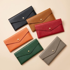 May include: Six rectangular envelope-style wallets with a textured surface, each featuring a single brass-colored snap button closure. The wallets are arranged in a cluster and come in black, tan, red, orange, dark green, and gray color options.