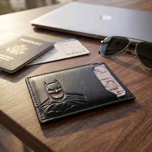 Könnte beinhalten: Ein schwarzer Kartenhalter aus Leder mit einem geprägten Batman-Logo auf der Vorderseite. Der Kartenhalter enthält einen sichtbaren Ausweis mit Porträt und Text. Der Artikel ist rechteckig mit genähten Kanten und einem flachen Design.