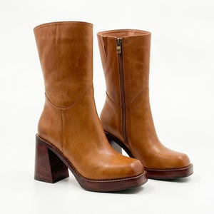 May include: A pair of mid-calf boots in a tan leather finish featuring a square toe, a side zipper closure, and a thick, dark brown wood-grain block heel and platform sole. The boots have a smooth, structured shaft and a clean, polished appearance.