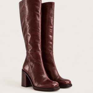 May include: A pair of knee-high boots in a deep burgundy color. These boots feature a smooth finish, a square toe, and a thick, block-style heel. The shaft of the boot reaches the knee, and the construction includes visible vertical seams.