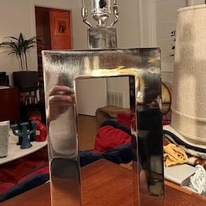 May include: A table lamp featuring a rectangular, polished silver-tone metal frame with a hollow center. The lamp sits on a solid, rectangular black base and includes a metal harp and socket assembly at the top for attaching a lampshade.