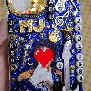 Peut inclure: Une coque de téléphone transparente décorée de paillettes bleues, une image centrale d'une personne en veste militaire bleue, un cœur rouge sur le visage, et divers charms dont des guitares, une couronne, un micro et des perles lettres MICHAEL JACKSON.
