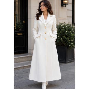 May include: A long, floor-length white coat featuring a structured collar, long sleeves, and a front button closure with three prominent gold-toned buttons. The coat has a tailored fit through the bodice and two side pockets with flap details.