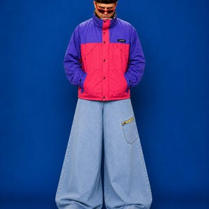 Puede incluir: Una chaqueta de bloques de color con mangas moradas y torso rosa brillante con una etiqueta negra, combinada con pantalones vaqueros de pierna ancha en azul claro con un bolsillo con cremallera en el muslo izquierdo y zapatillas blancas.