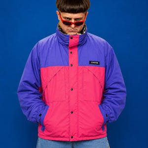 May include: A color-blocked puffer jacket featuring a vibrant purple upper section and a bright pink lower section. The jacket has a high collar, a front snap-button closure, and a black rectangular patch on the chest with the word CUSTOM in white.