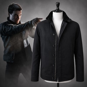 Puede incluir: Una chaqueta negra expuesta en un maniquí, con cuello vuelto con forro interior oscuro, cierre central de cremallera y una hilera vertical de cinco botones negros a juego. La chaqueta tiene mangas largas con puños y dos bolsillos laterales.