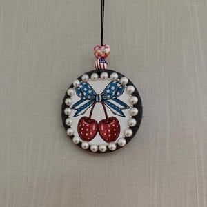 May include: A circular hanging ornament featuring a central graphic of two red cherries with white stars and a blue and white striped bow. The edge is lined with white pearls. Above the ornament are two beads, one checkered and one with a flag pattern.