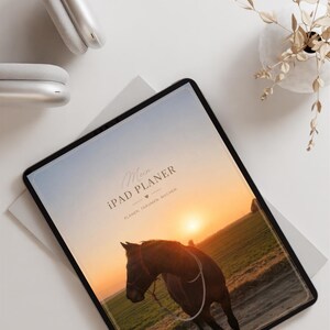 Könnte beinhalten: Ein digitales Tablet mit einem Planer-Cover, das den Text Mein IPAD PLANER PLANEN TRAUMEN MACHEN und ein Bild eines braunen Pferdes auf einem Feld bei Sonnenuntergang zeigt, neben weißen Over-Ear-Kopfhörern und einer kleinen Vase mit Trockenblumen.