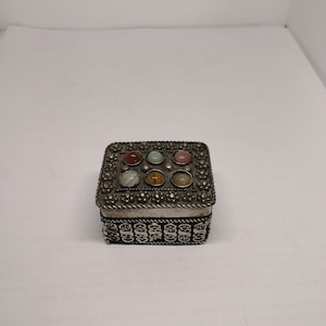 Può includere: Una scatola quadrata in metallo con finitura color argento ossidato, caratterizzata da intricati motivi floreali in rilievo lungo i bordi. Il coperchio è decorato con sei pietre cabochon lisce, rotonde e multicolori disposte in due file.