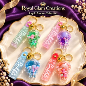 May include: Four astronaut-shaped keychains with liquid-filled domes containing colorful beads. Each keychain features a rectangular silicone strap with the word Bubble printed in white, a gold-tone metal ring, and a small gold bell charm.