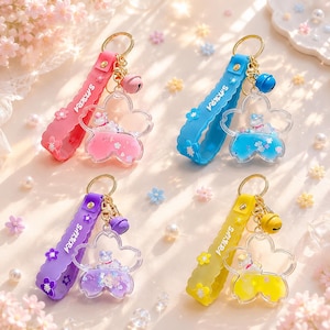 May include: Four keychains in pink, blue, purple, and yellow. Each features a silicone strap with the word SAKURA, a small bell, and a transparent flower-shaped charm containing liquid, glitter, and a small white animal figure inside.