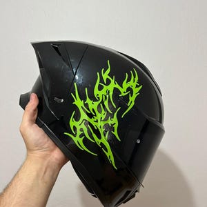 Peut inclure: Un casque de moto intégral noir brillant orné d'un motif de flammes tribales vert néon sur le côté. Le casque est doté d'une visière teintée foncée, d'une coque aérodynamique profilée et d'un aileron arrière pour une meilleure stabilité.