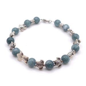 Könnte beinhalten: Ein Perlenarmband mit runden, blau-grünen Steinperlen, die mit silberfarbenen Metallanhängern in Form von Meeresschildkröten und einem Fischschwanz durchsetzt sind. Das Armband wird mit einem silberfarbenen Karabinerverschluss geschlossen.