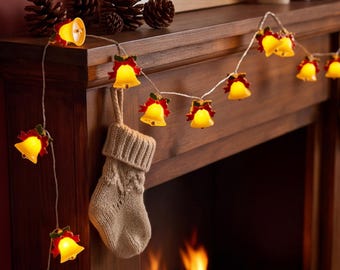 Puede incluir: Una guirnalda de luces con forma de campana, cada una con una campana amarilla y un lazo rojo. Las luces están dispuestas sobre una repisa de madera junto a un calcetín de punto beige y varias piñas de color marrón.
