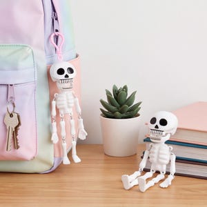 May include: Two white plastic skeleton figures with black eye sockets and teeth. One skeleton is attached to a pastel rainbow backpack by a pink plastic clip, while the other sits on a wooden surface next to a small green succulent in a white pot.