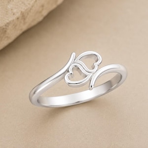 May include: A polished silver-toned ring featuring an open bypass design with two interconnected heart-shaped outlines. The band is smooth and rounded, tapering slightly as it curves to form the central heart motif, creating a continuous loop.
