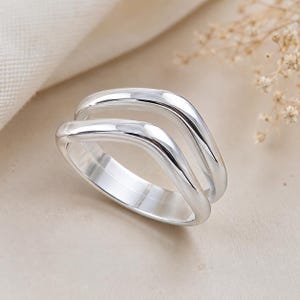 Puede incluir: Un anillo de plata pulida con un diseño de doble banda y una forma ondulada suave y continua. El metal tiene un acabado reflectante de alto brillo, creando una apariencia elegante y uniforme en las dos bandas curvas interconectadas.