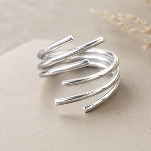 Può includere: Un anello in argento lucido caratterizzato da un design a spirale multistrato. La fascia è composta da diverse bobine lisce e arrotondate che si avvolgono attorno al dito, creando una struttura aperta e sovrapposta con finitura metallica.