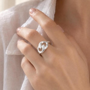 May include: A polished silver ring featuring an interlocking, abstract knot design. The band consists of two smooth, rounded metal loops that overlap to create a continuous, fluid shape. The surface of the silver is reflective and bright.