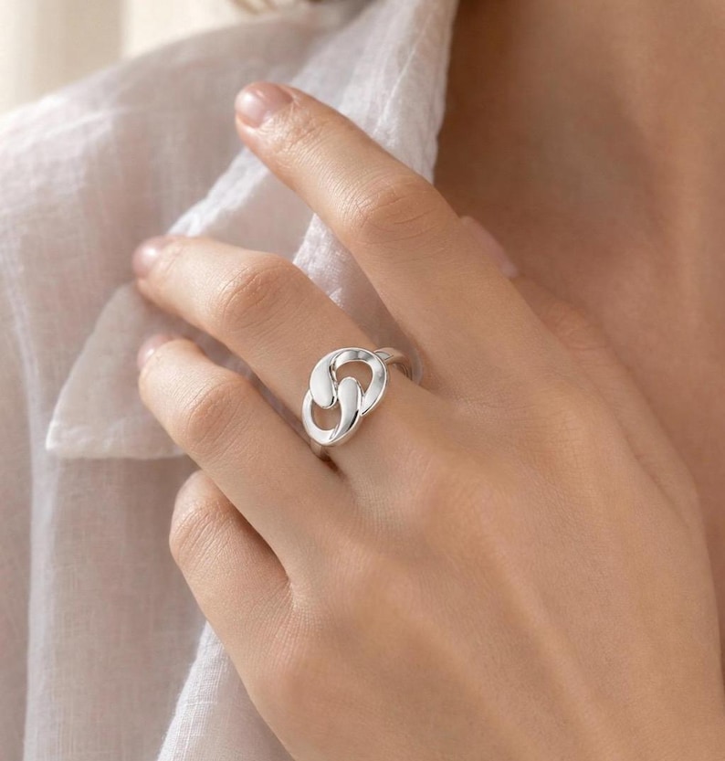 May include: A polished silver ring featuring an interlocking, abstract knot design. The band consists of two smooth, rounded metal loops that overlap to create a continuous, fluid shape. The surface of the silver is reflective and bright.