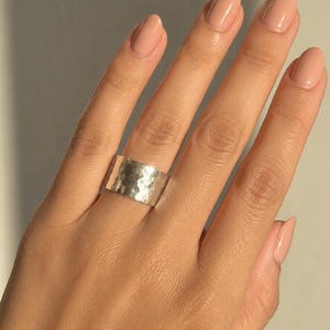 May include: A wide silver band ring featuring a textured, hammered finish across its entire outer surface. The metal has a reflective, polished appearance with numerous small, irregular indentations that create a light-catching effect.
