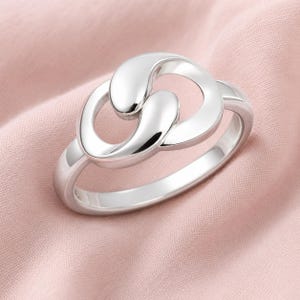 May include: A polished silver ring featuring a central design of two interlocking, smooth, teardrop-shaped loops. The metal has a reflective, high-shine finish, and the band is a simple, solid, rounded silver hoop that connects to the focal point.
