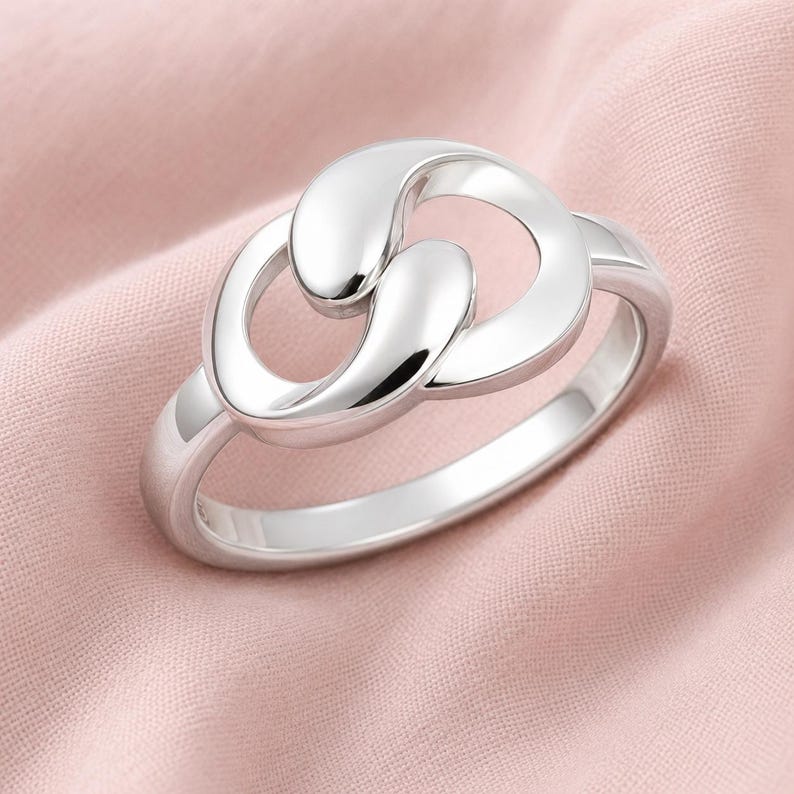May include: A polished silver ring featuring a central design of two interlocking, smooth, teardrop-shaped loops. The metal has a reflective, high-shine finish, and the band is a simple, solid, rounded silver hoop that connects to the focal point.