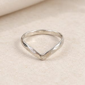 Peut inclure: Une bague de couleur argentée avec un design en forme de V ou de chevron. L'anneau présente une finition martelée texturée sur toute sa surface, créant une série de petites facettes réfléchissantes qui captent la lumière au centre.
