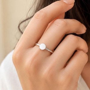 Op de afbeelding: Een gepolijste zilverkleurige ring met een glad, plat, cirkelvormig schijfje bevestigd aan een dunne, afgeronde band. Het metalen oppervlak is reflecterend en heeft een uniforme kleur, wat een eenvoudig en strak ontwerp zonder extra texturen of patronen laat zien.