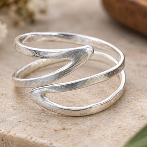 Puede incluir: Un anillo de plata con un diseño de bypass con dos bandas abiertas que se superponen. La superficie del metal tiene un acabado martillado y texturizado, creando una apariencia reflectante. El anillo está hecho de una pieza de metal plateado.