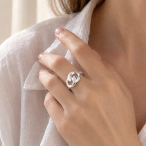 May include: A polished silver ring featuring an interlocking double-loop design. The ring has a smooth, reflective metallic surface and a continuous, fluid shape that wraps around the finger, creating a modern and clean aesthetic appearance.