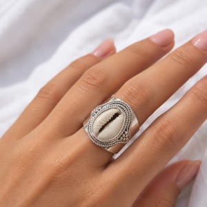 May include: A wide silver band ring featuring a central oval cowrie shell. The shell is framed by a decorative twisted rope border and small silver bead accents. The metal band has a polished finish and a wide, flat profile that tapers slightly.