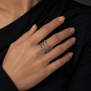 Könnte beinhalten: Ein silberfarbener Ring mit einem dreifachen Banddesign, das in der Mitte einen ausgeprägten V-förmigen Winkel aufweist. Das Metall hat eine glatte, polierte Oberfläche, wobei die drei Bänder zu einem einzigen Schmuckstück verbunden sind.