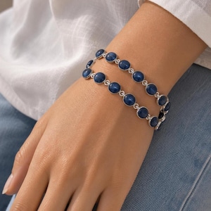 Puede incluir: Una pulsera de doble cadena con piedras cabujón redondas de color azul intenso engastadas en monturas de metal plateado pulido. Las piedras están unidas en un diseño de cadena continua que rodea la muñeca, creando un aspecto superpuesto.