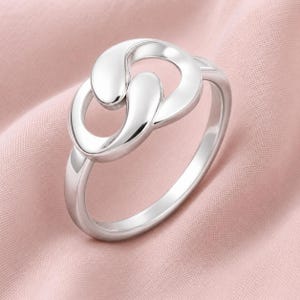 May include: A polished silver ring featuring a central design composed of two interlocking, smooth, teardrop-shaped loops. The metal has a bright, reflective finish, and the band is a simple, solid circular shape that connects to the focal point.