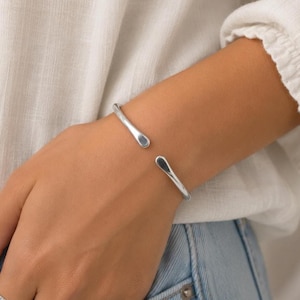 May include: A polished silver-tone cuff bracelet with a smooth, open-ended design. The bracelet features two rounded, tapered ends that face each other, creating a simple, continuous metallic band that rests comfortably around the wrist area.