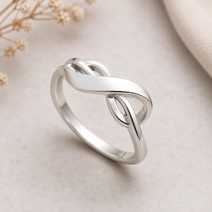 Può includere: Un anello color argento lucido caratterizzato da un design a simbolo di infinito continuo. La fascia è liscia e riflettente, mostrando una forma a intreccio fluido che costituisce l'elemento centrale del gioiello, creando un aspetto pulito.