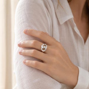 May include: A polished silver ring featuring an interlocking double-link design. The band consists of two smooth, rounded metal loops that are joined together in a continuous, fluid shape, resting on the ring finger of a hand.