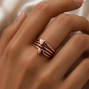 May include: A rose gold ring featuring a multi-band design with two small, polished heart-shaped charms attached to the bands. The metal has a smooth, reflective finish, and the ring is constructed from several thin, stacked circular bands.