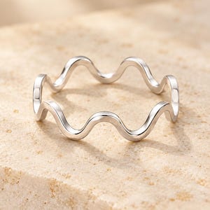 Puede incluir: Un anillo de tono plateado pulido que presenta un diseño de banda continua ondulada o serpentina. La superficie metálica es lisa y reflectante, creando una serie de picos y valles uniformes que forman la silueta circular de la pieza.