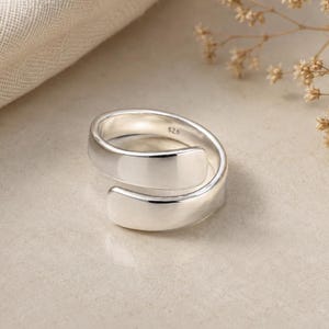 Puede incluir: Un anillo de plata pulida con diseño abierto tipo bypass. La banda interior está claramente marcada con el sello S925, que indica su composición. El anillo presenta un acabado metálico reflectante con bordes redondeados y un bucle continuo.