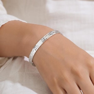 Op de afbeelding: Een zilverkleurige metalen armband met een getextureerd, gehamerd oppervlak. De armband heeft een slank, open ontwerp dat om de pols valt en een reflecterende, metalen glans vertoont over de gehele buitenste omtrek van het sieraad.