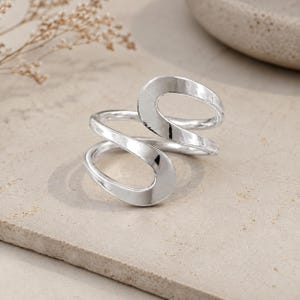 Può includere: Un anello in argento lucido caratterizzato da un design aperto a bypass con due fasce curve sovrapposte che creano una forma fluida e continua. La superficie metallica è liscia e riflettente, con una finitura brillante su tutto il pezzo.