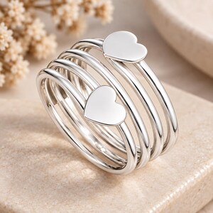 May include: A silver-toned multi-strand ring featuring a coiled design with two flat heart-shaped accents at the ends. The ring is constructed from several smooth, polished metal bands that wrap around to create a layered, continuous appearance.