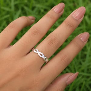 Op de afbeelding: Een zilveren ring met een doorlopend gedraaid bandontwerp in infinity-stijl. Het metaal heeft een gladde, gepolijste afwerking en de band bestaat uit herhalende ovaalvormige schakels die een delicaat, onderling verbonden patroon vormen.